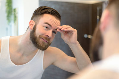 Reflection of young man tweezing eyebrows on mirror in bathroom