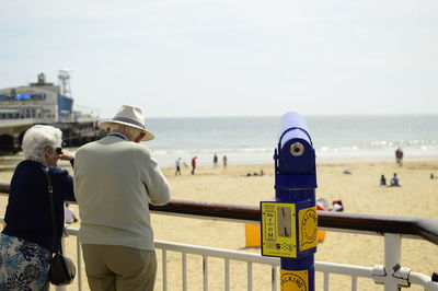 Senior couple standing by coin-operated binoculars at beach on sunny day