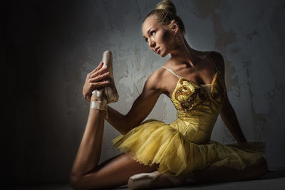 Beautiful ballerina stretching while sitting against wall in dark
