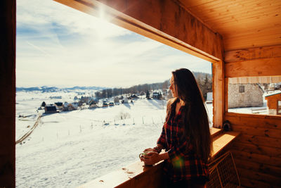 Young woman holding cup while standing by window in cottage during winter