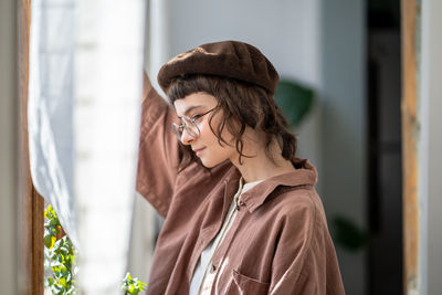 Thoughtful teenager standing at window at home alone, staring attentively at street, waiting