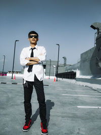 Full length of young man with arms crossed standing on walkway against clear sky