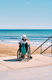 Back view of unrecognizable female traveler in wheelchair with backpack enjoying summer journey on beach near blue sea