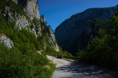 Narrow road along trees and mountains against clear sky