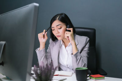 Businesswoman talking on mobile phone while sitting at desk against wall in office