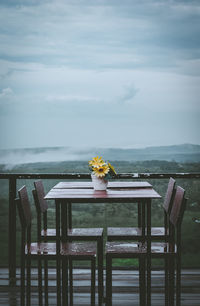 Empty chairs with table by railing against sky