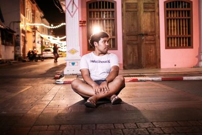 Young man looking away while sitting on footpath in city