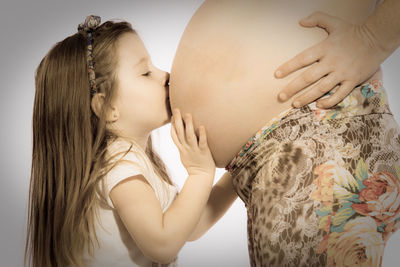 Side view of girl kissing pregnant mother while standing against white background