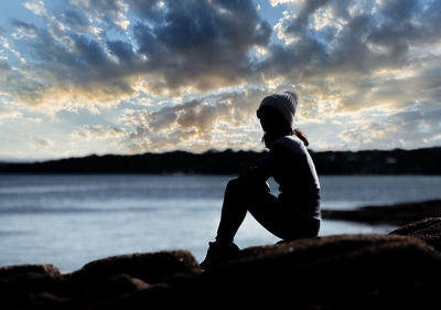 Man sitting on rock by sea against sky during sunset