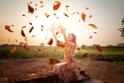 Smiling young woman throwing dry leaves while sitting against sky during sunset