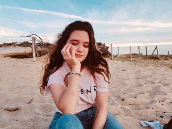 Beautiful young woman sitting on sand at beach against sky