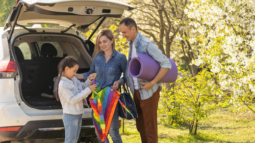 Happy family standing together near a car with open trunk enjoying view of rural landscape nature. 