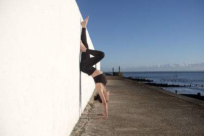 Sporty woman doing a handstand against a white wall by the sea. outdoor exercise concept.