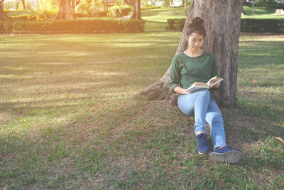 Full length of young woman reading book against tree at park