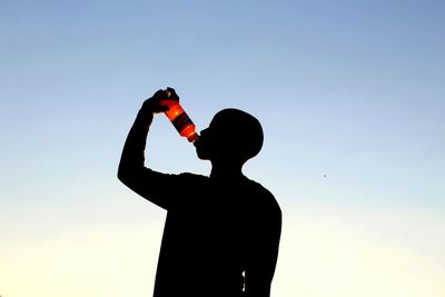 Low angle view of silhouette man drinking while standing against clear sky during sunset