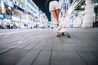 Low section of woman skateboarding on footpath in illuminated city during night