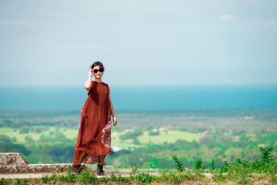 Portrait of young woman wearing sunglasses standing on observation point against cloudy sky