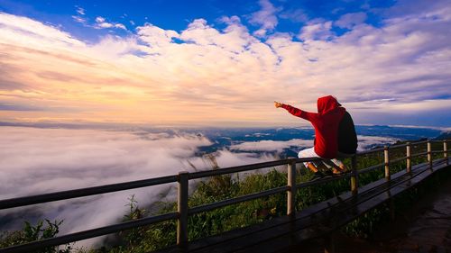 Woman and man sitting on railing against sky during sunset