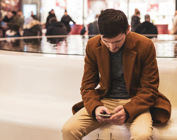 A young man with a phone in his hands is sitting on a bench in a store.