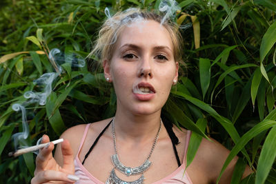 Portrait of young woman smoking marijuana joint while standing against plants