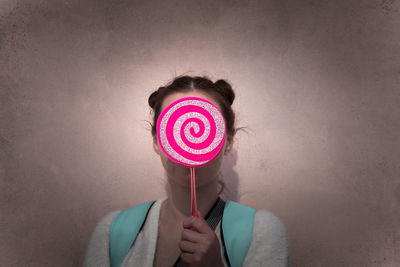 Woman covering face with pink lollipop against wall