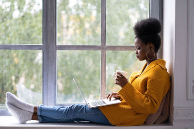 Side view of woman using laptop while holding coffee by window