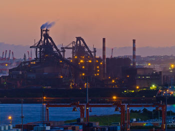 Illuminated cranes at harbor against sky during sunset