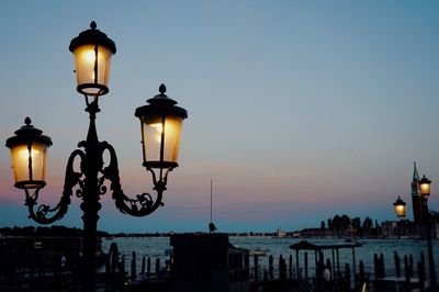 Illuminated street light by sea against sky during sunset