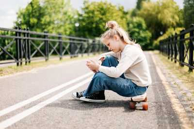 Cute blonde teenage girl sitting on a skateboard in a city park chatting on a smartphone
