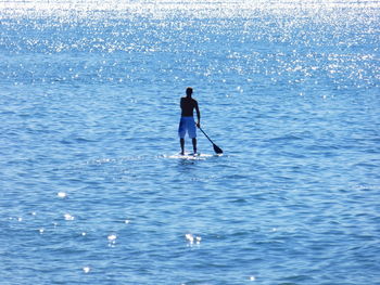 Full length rear view of shirtless man paddleboarding on sea during sunny day