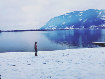 Woman standing on snowcapped land by lake against sky