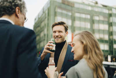 Smiling male entrepreneur with drink looking at coworkers in city