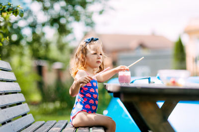 A cute baby is drinking a berry smoothie through a straw, sitting on a bench in the garden. 