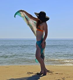 Full length side view of sensuous young woman holding sarong at beach against clear blue sky
