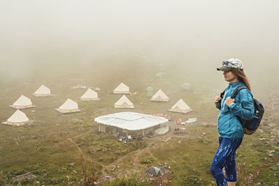 Young woman in cap blue sportswear with backpack hiker  in mountain  against tent camping in fog