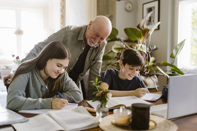 Smiling grandfather looking at grandchildren doing homework while sitting at table in home