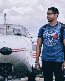 Young man looking away while standing by airplane against cloudy sky