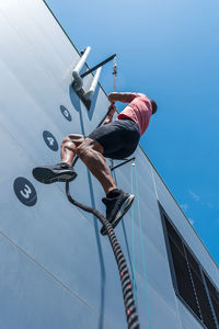 From below of confident male athlete climbing rope while doing exercises on sports ground in summer on sunny day