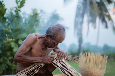 Shirtless man making wicker baskets against trees