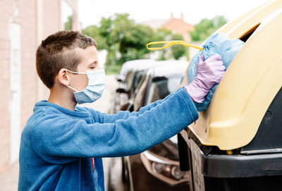 Boy throwing a blue trash bag into a yellow recycling container in the street. the kid wearing face mask and gloves is pushing the rubbish bag because it is too big to enter the bin. horizont