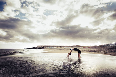 Side view of young woman practicing yoga in bridge position at beach against cloudy sky