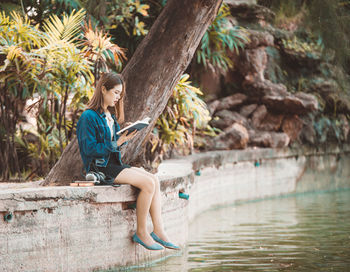 Full length of young woman reading book while sitting by lake