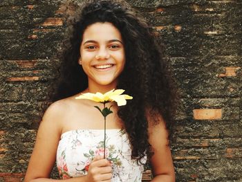 Portrait of smiling teenage girl with artificial flower standing against brick wall