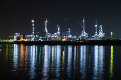 Illuminated factory by river against sky at night