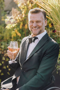 Portrait of happy man wearing suit holding wineglass sitting at wedding