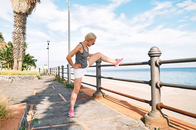 Young beautiful sportive woman training over seaside promenade, stretching legs before jogging.