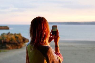 Rear view of woman photographing sea against sky during sunset