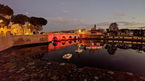 Illuminated buildings by lake against sky at dusk