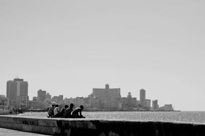 Friends sitting on retaining wall by sea against clear sky