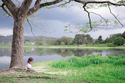 Girl sitting under tree trunk by lake against sky